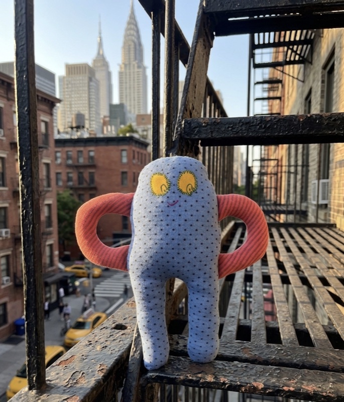 Handmade light-blue polka-dot stuffed creature with long orange striped looping arms and yellow embroidered eyes, posed on a fire escape with the New York City skyline behind it