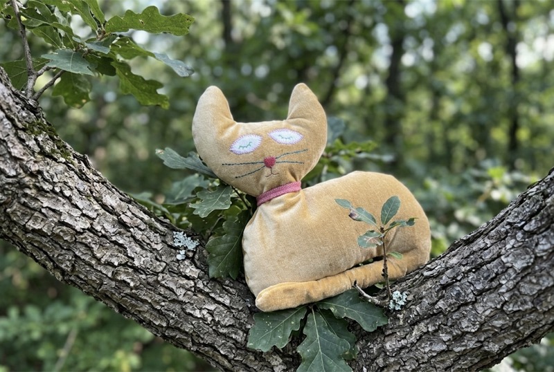 Handmade stuffed cat in golden fabric with embroidered sleeping eyes and pink nose, perched in an oak tree