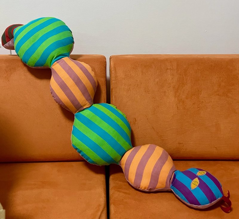 Striped caterpillar pillow: a long handmade stuffed caterpillar in green-and-teal and orange-and-mauve striped segments, with a purple and teal striped head, appliqué leaf eyes and a little tongue, stretched across an orange sofa
